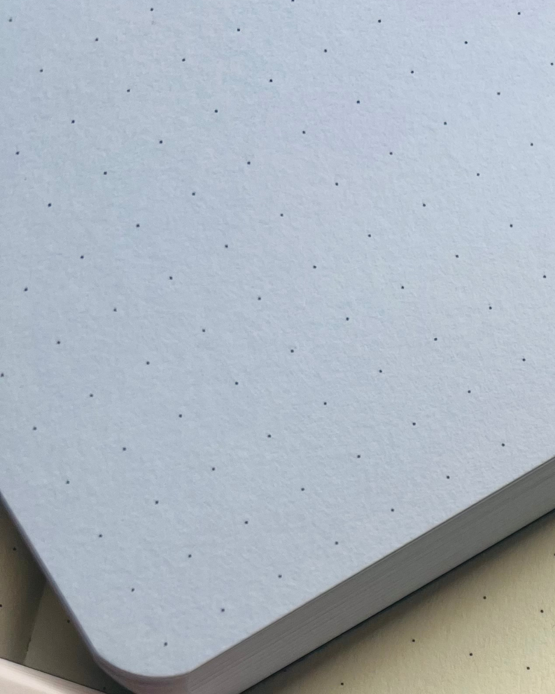 A close-up of a light blue-gray dotted notebook page with a textured paper surface and a stack of similar pages visible along the edge.