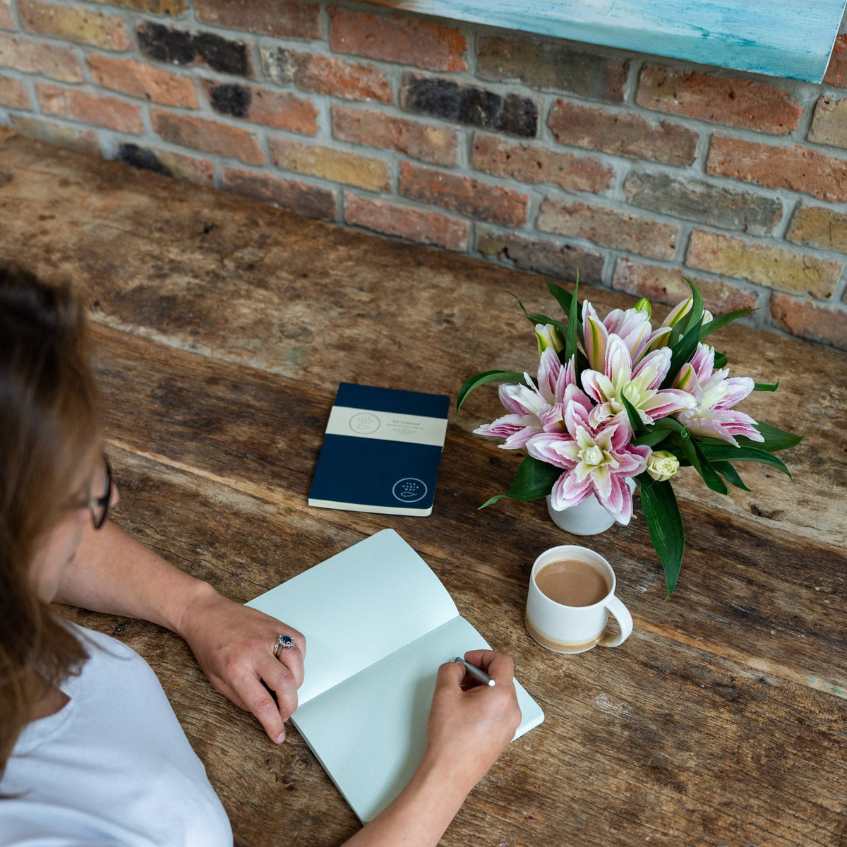 Person writing in a notebook on a wooden desk with a coffee cup and flowers, against a brick wall.